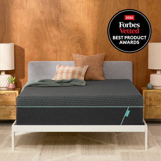 A Tuft and Needle Mint Hybrid mattress on a white bed frame in a bedroom setting with a wood-paneled wall, bedside tables, lamps, and two pillows. A circular 'Forbes Vetted 2026 Best Product Awards' badge is in the top right corner.