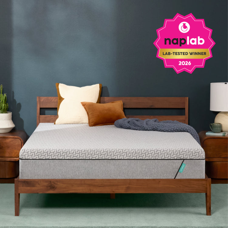 A Tuft and Needle Mint mattress on a wooden bed frame in a styled bedroom setting with a dark blue wall, bedside tables, pillows, and a pink NapLabs award badge in the top right corner.