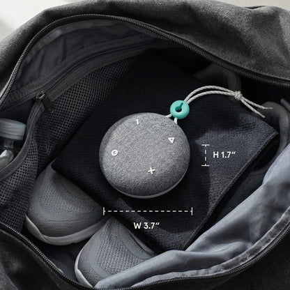A Tuft and Needle SNOOZ Go 2 Travel White Noise Sound Machine, round gray fabric speaker, with white control symbols and a small teal hanging ring, shown inside an open black bag with size annotations reading 3.7 inches wide, 1.