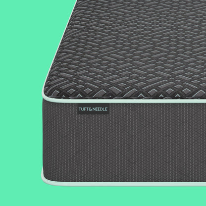 Close-up corner view of a the Tuft and Needle 15 inch Mint Luxe Hybrid Plush mattress showing the dark textured top surface and side panel with a visible TUFT & NEEDLE label, set against a solid mint green background.