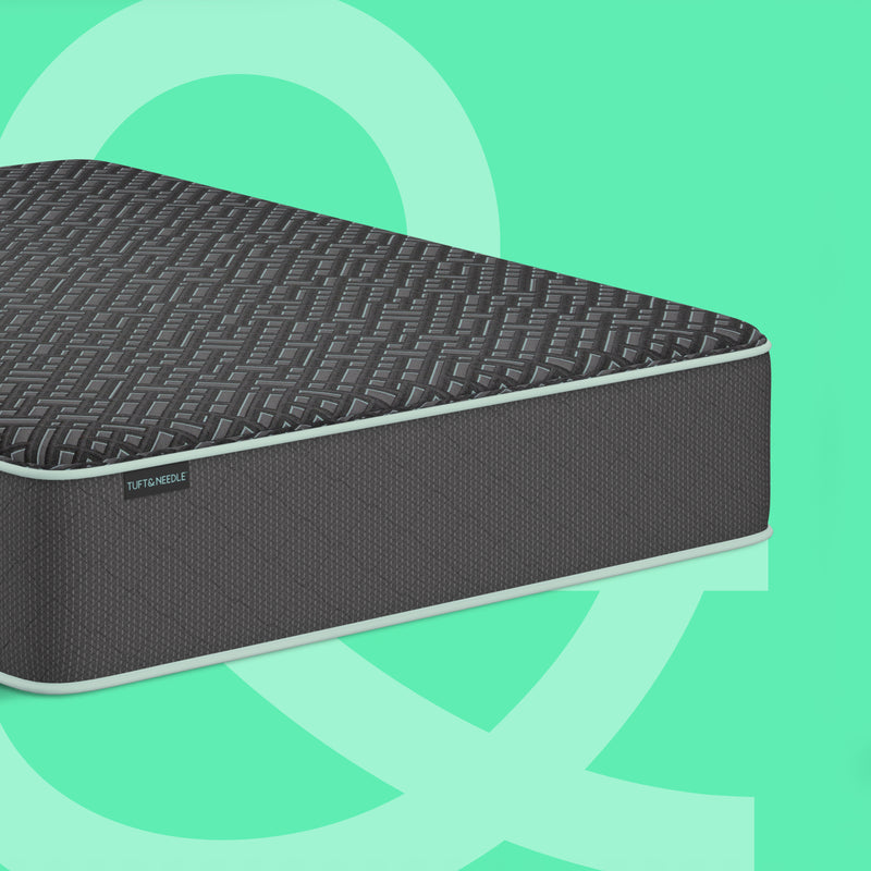 A dark gray Tuft & Needle 14.5 inch Mint Luxe Hybrid Medium textured mattress shown at an angle against a mint green background with abstract light green shapes. The brand name Tuft & Needle is visible on a small label on the side of the mattress.