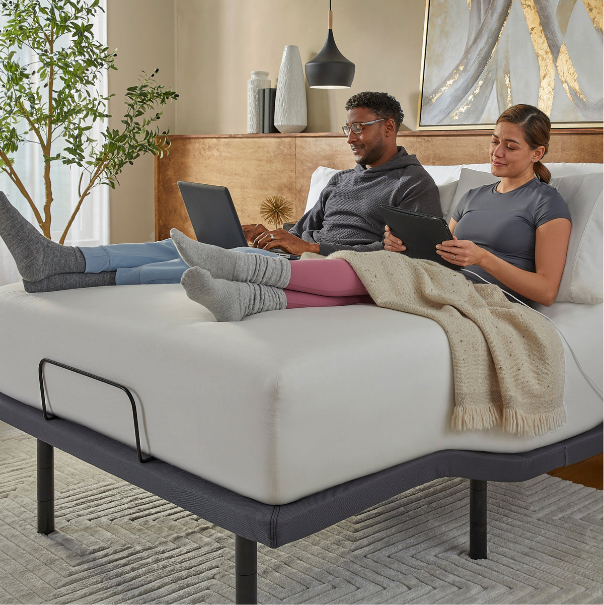 Two people using laptops on a mattress on top of a Baselogic Pearl adjustable base in a cozy room.
